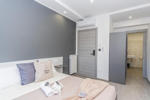 A modern hotel room with a bed, a radiator, and a bathroom visible through an open door. - Bisuite Rooms (Naples)
