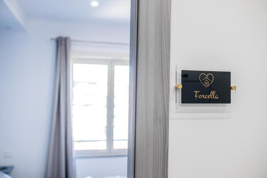 Une chambre d’hôtel avec un lit, une fenêtre avec des rideaux et une enseigne fixée au mur portant le nom « Forcella ».