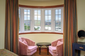 A room with a bay window, two pink chairs, a small round table, and a mirror on a blue dresser.