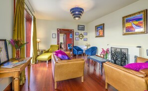 A living room with a wooden floor, a sofa, a coffee table, a blue armchair, and a yellow armchair. There is a fireplace with a decorative mirror and a framed picture on the wall. - Oban Bay Hall - Seven Bedroom Villa, Sleeps 14 (Oban)