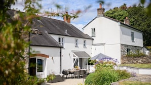 A white building with a stone facade, a covered patio with outdoor seating, and a garden with various plants.