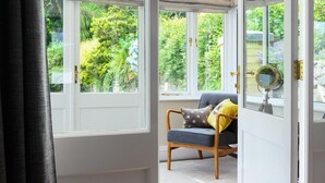 A conservatory with a view of a lush garden, a wooden chair with cushions, and a round mirror on a stand.