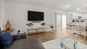 A modern living room with a flat-screen TV, a sofa, a coffee table, and a dining area with a wooden table and white chairs. - Cozy 2BR Near Eastport Sq. & Middleton | Patio (Sumterville)