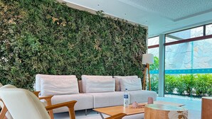 A modern hotel room with a green leafy wall, a sofa, a wooden coffee table, and a lamp.