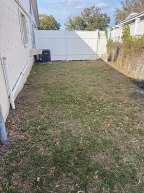 A backyard with a white fence, a lawn, and a building with a window and a vent.