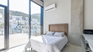 Ein Schlafzimmer mit Bett, Schreibtisch und Blick auf eine Stadtlandschaft durch große Fenster.