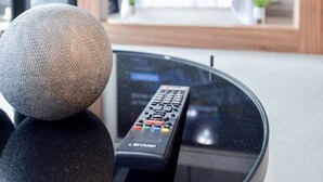 Ett modernt vardagsrum med ett soffbord i glas, en fjärrkontroll och en stor tv-skärm som hänger på väggen och visar flera kanaler.