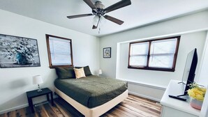 A bedroom with a bed, a ceiling fan, a desk with a computer, and two windows with blinds.