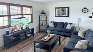 A modern living room with a dark grey sofa, a wooden coffee table, and a TV on a black stand. There is a bookshelf with books and decorative items, a mirror on the wall, and a clock.