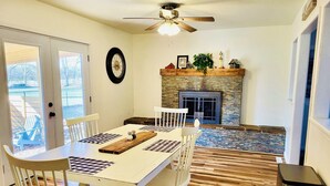A dining area with a white table, chairs, and a wooden floor. There is a fireplace with a stone surround and a ceiling fan above.