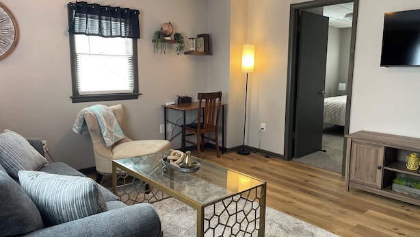 Una sala de estar con sofá, mesa de centro de vidrio, una silla, lámpara y televisor.