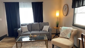 Una sala de estar con un sofá gris, un sillón beige, una mesa de centro de vidrio y una mesita auxiliar con una lámpara.