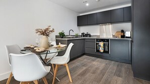 Uma cozinha moderna com armários escuros, uma mesa de jantar de vidro, cadeiras brancas e um vaso com flores secas.