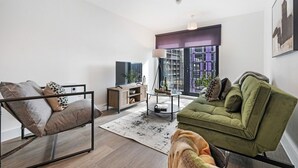 Uma sala de estar moderna com um sofá verde, uma poltrona de metal, uma mesa de centro de vidro e uma grande janela com vista para a cidade.