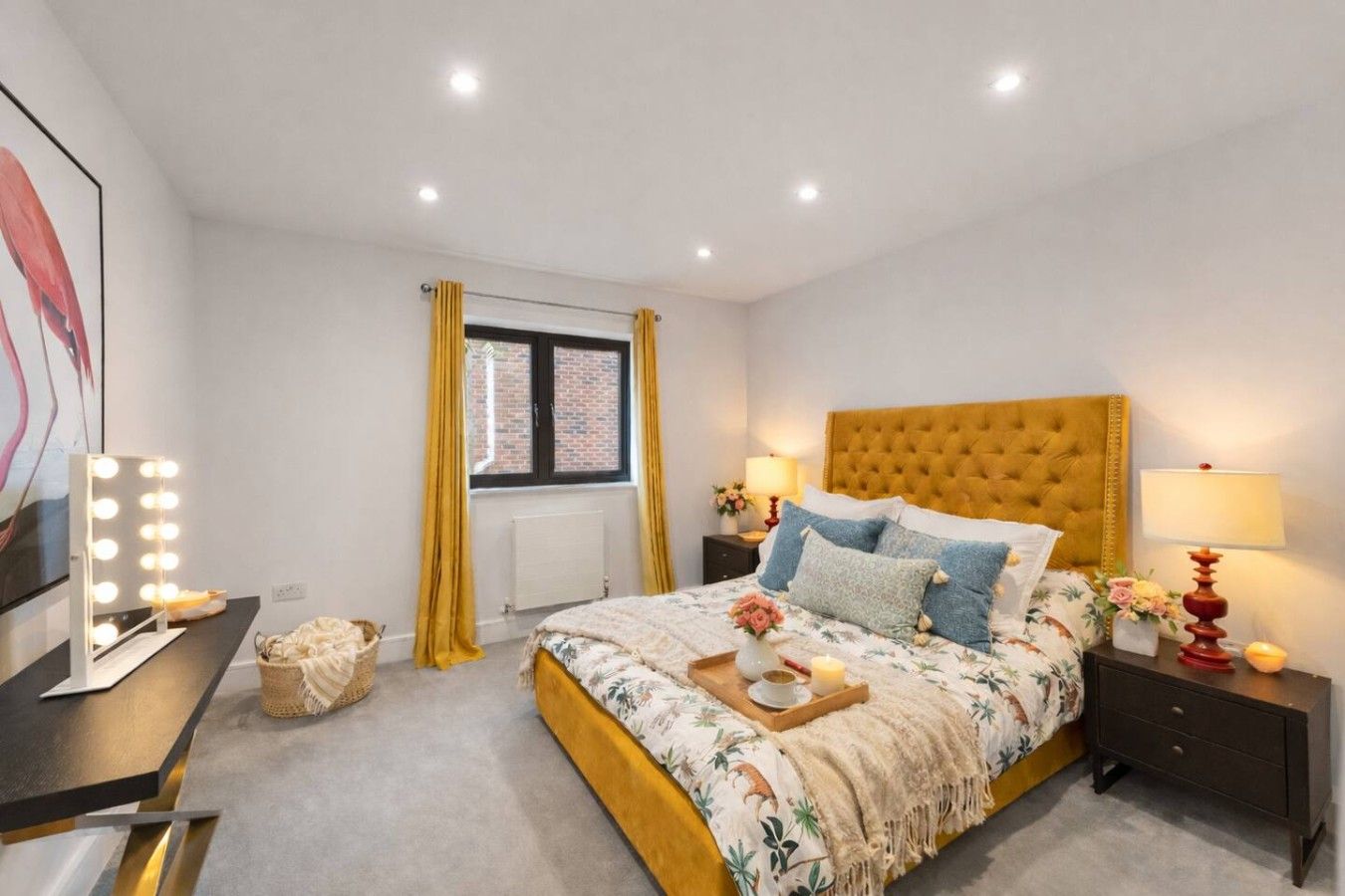 A bedroom with a bed, bedside tables, a vanity with lights, a window with curtains, and a wall-mounted mirror.