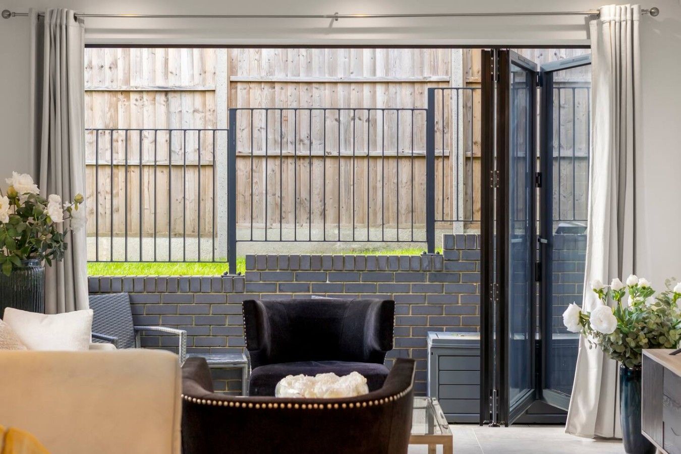 A room with a sliding glass door leading to a fenced outdoor area, a black chair, a sofa, and a vase with flowers.