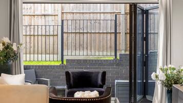 A room with a sliding glass door leading to a fenced outdoor area, a black chair, a sofa, and a vase with flowers.