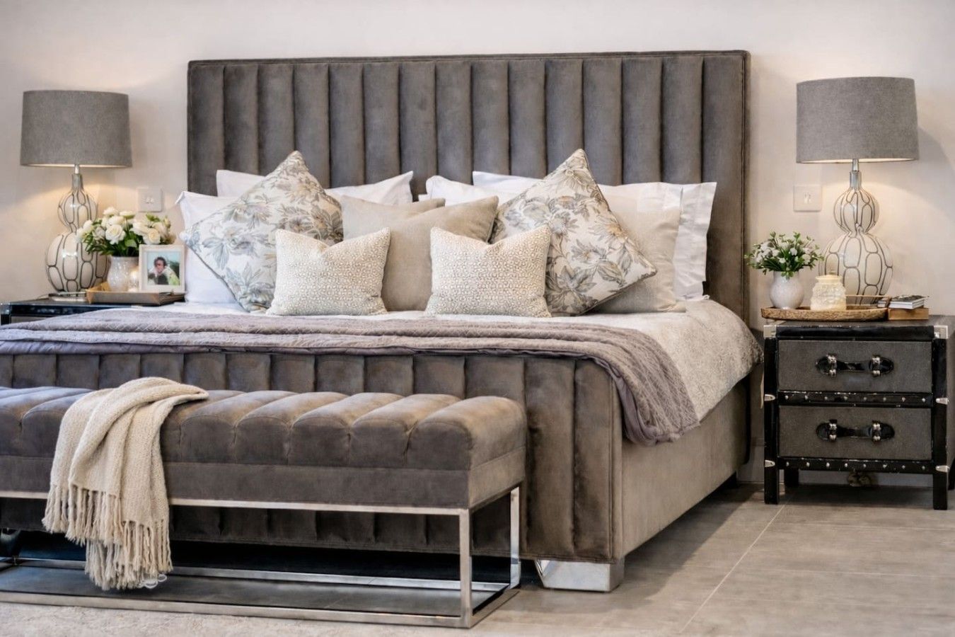 Un dormitorio con cabecera capitonada, una cama grande con varias almohadas, un banco al pie de la cama, una mesita de noche con lámpara y un aparador con cajones.