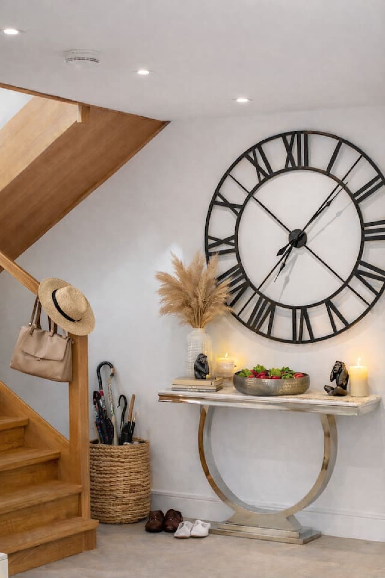 A staircase with a wooden finish, a large wall-mounted clock, a small table with decorative items, and a wicker basket with shoes.