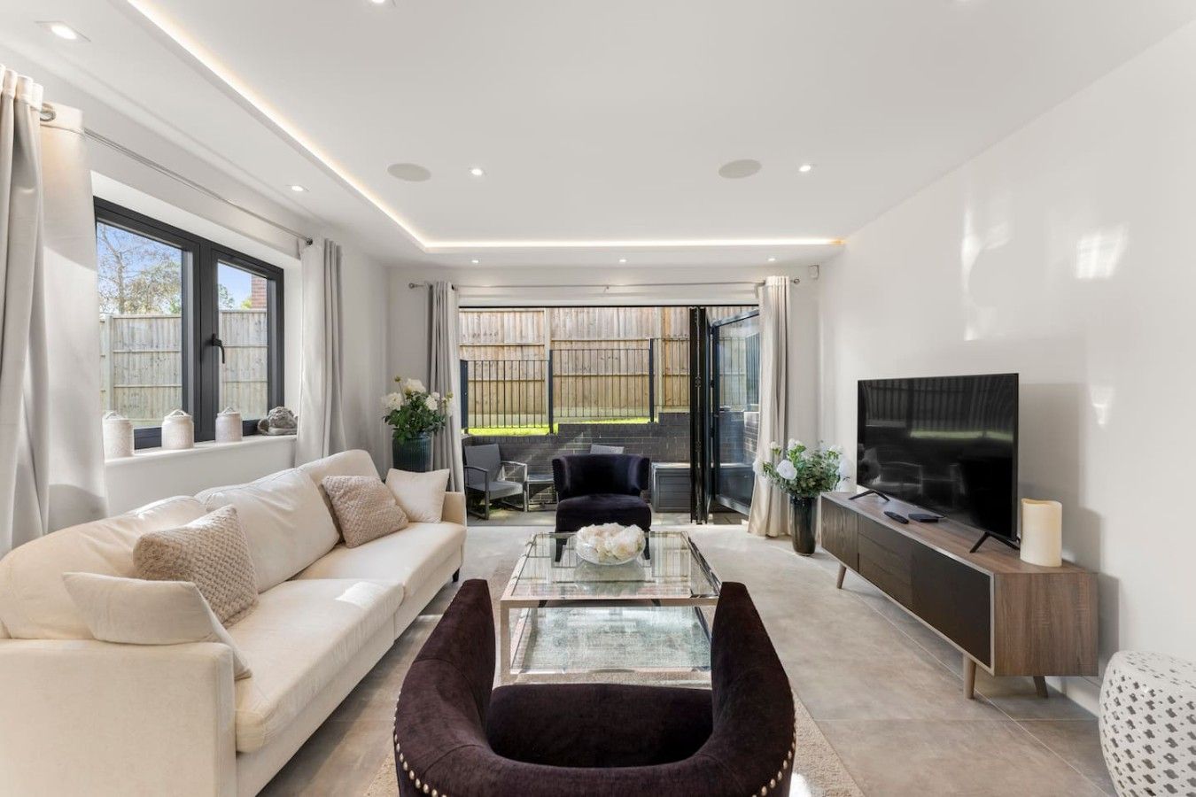 A modern living room with a sofa, armchair, and TV, featuring a glass coffee table and a view through the glass door.