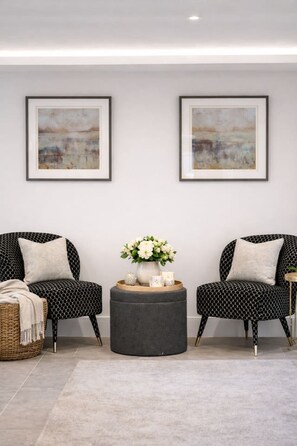 Ein Zimmer mit zwei schwarzen, gemusterten Stühlen, einem runden Hocker, einem Korpus-Beistelltisch und einer Vase mit Blumen.