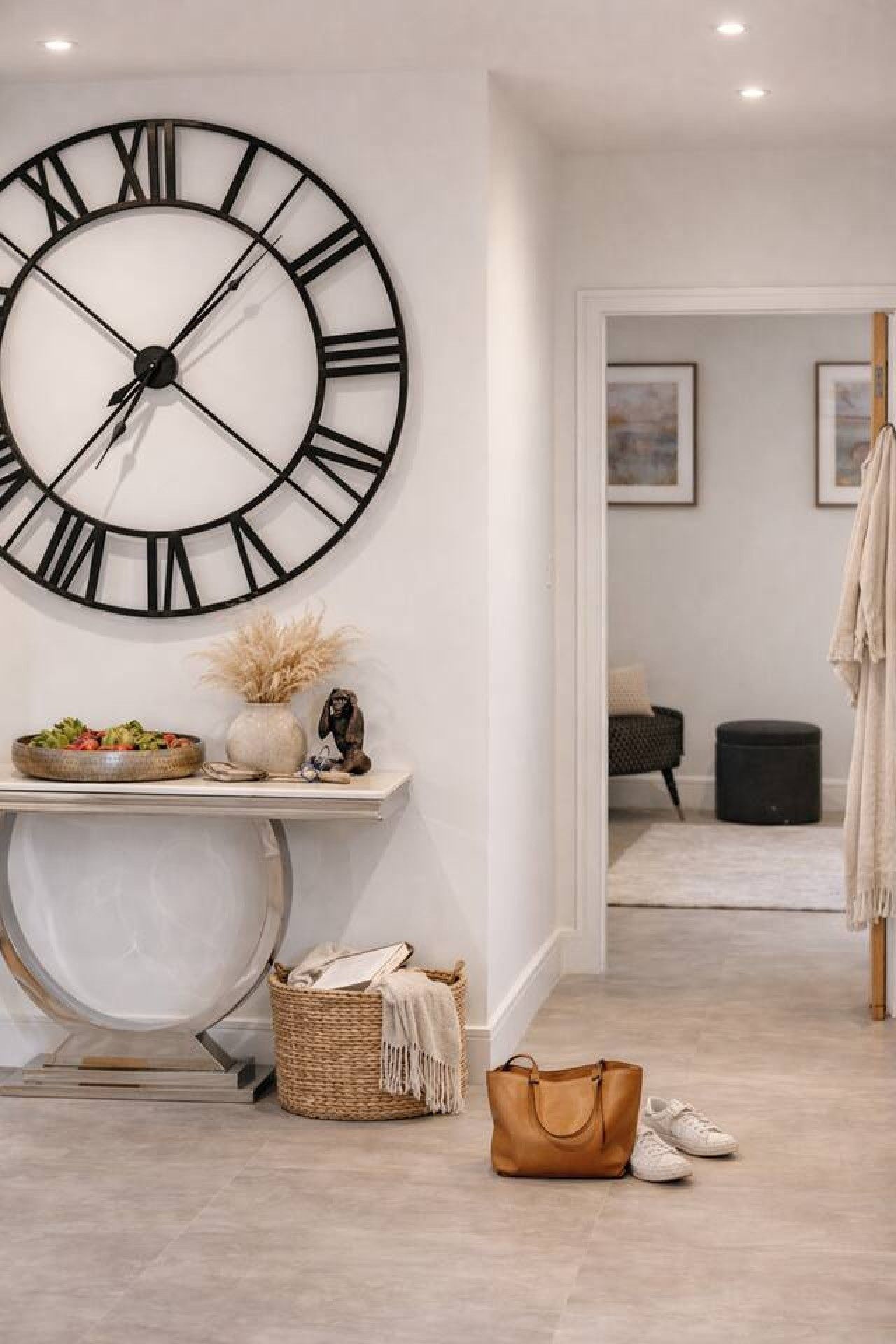 A hallway with a large wall clock, a console table with decorative items, a wicker basket, a brown handbag, and a pair of white sneakers.