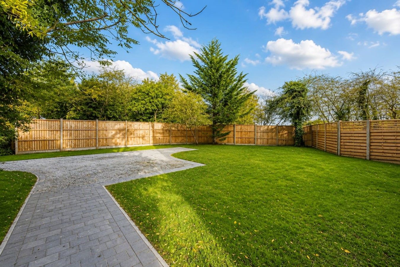 A backyard with a paved pathway, a grassy lawn, and a wooden fence.
