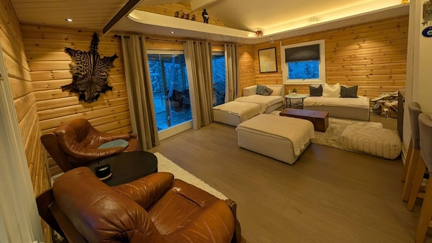 Un accogliente interno di una baita, con arredamento in legno, un divano in pelle, un tavolo da pranzo e un'ampia finestra che si affaccia sulla natura.