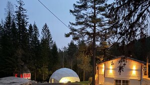 Una baita rustica con una struttura a cupola sullo sfondo, circondata da alberi e un sentiero sterrato.