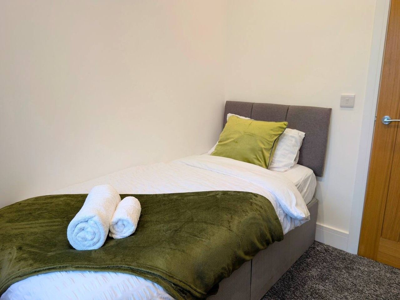 A single bed with a grey headboard, a green throw blanket, and two rolled white towels.