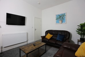 A living room with a leather sofa, a coffee table, a flat-screen TV, a radiator, and a painting on the wall. - Contractor House, 4 Bed Ensuite, Near Stadium (West Midlands)