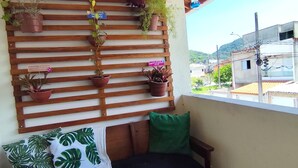 A wooden bench with a green cushion and a white throw blanket, a wooden slat wall with potted plants, and a view of buildings and hills in the background. - Cozy house in Guarujá, 3km from the beach. Air conditioning/Wi-Fi/barbecue. (Guarujá)
