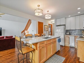 A modern kitchen with wooden cabinets, a central island, and pendant lighting. - Coastal House Near the Beach | Kitchen, Deck & BBQ (Bethany Beach)