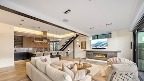 A modern living room with a large sofa, a coffee table, and a fireplace. There is a kitchen in the background with a staircase leading to an upper level. - Hot Tub with Slope Views | Walk to Lift (Park City)