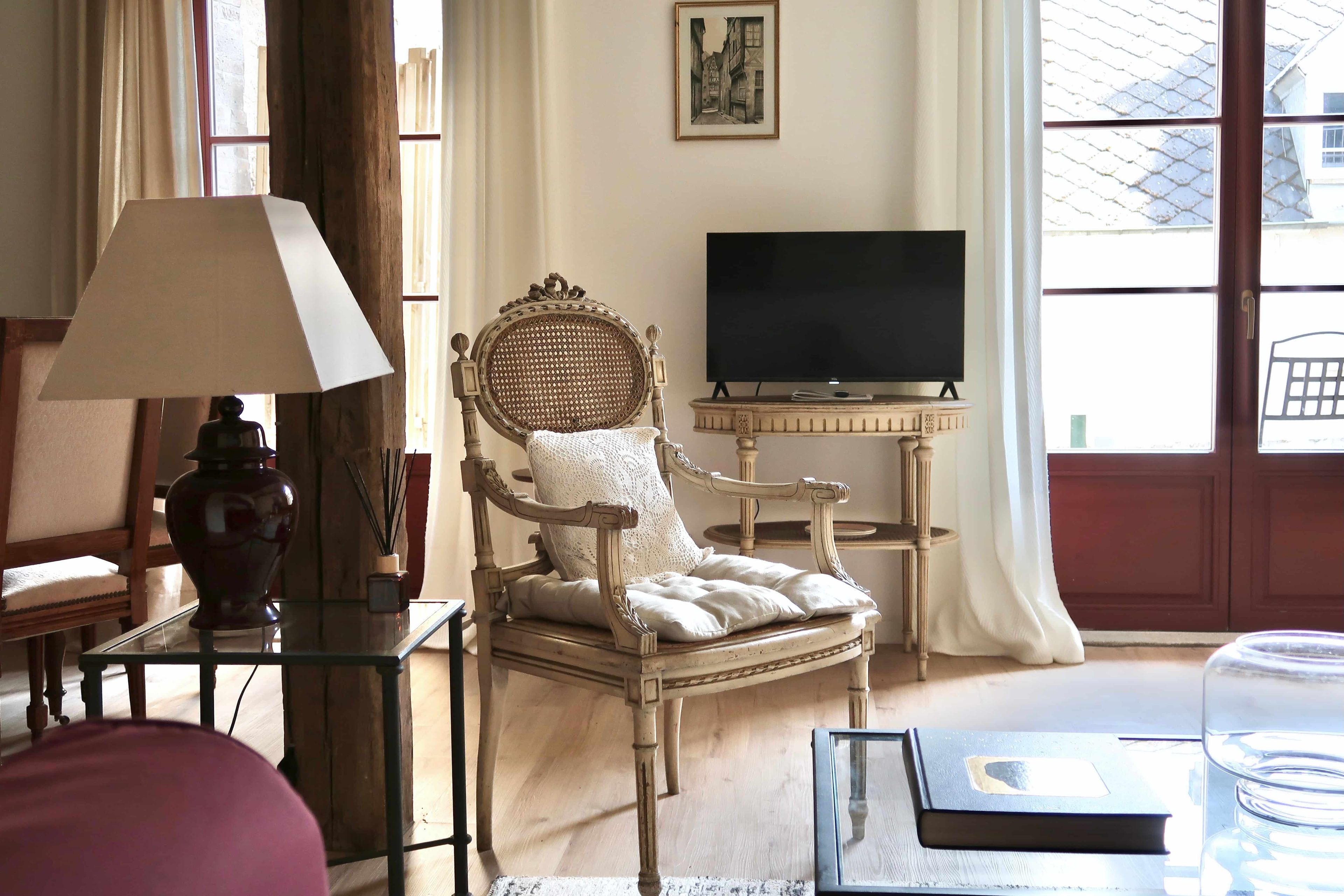 Une pièce avec une chaise en bois, une table en verre, un téléviseur et une fenêtre avec des rideaux.