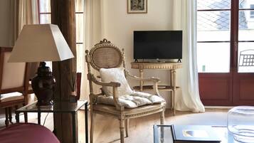 Une pièce avec une chaise en bois, une table en verre, un téléviseur et une fenêtre avec des rideaux.