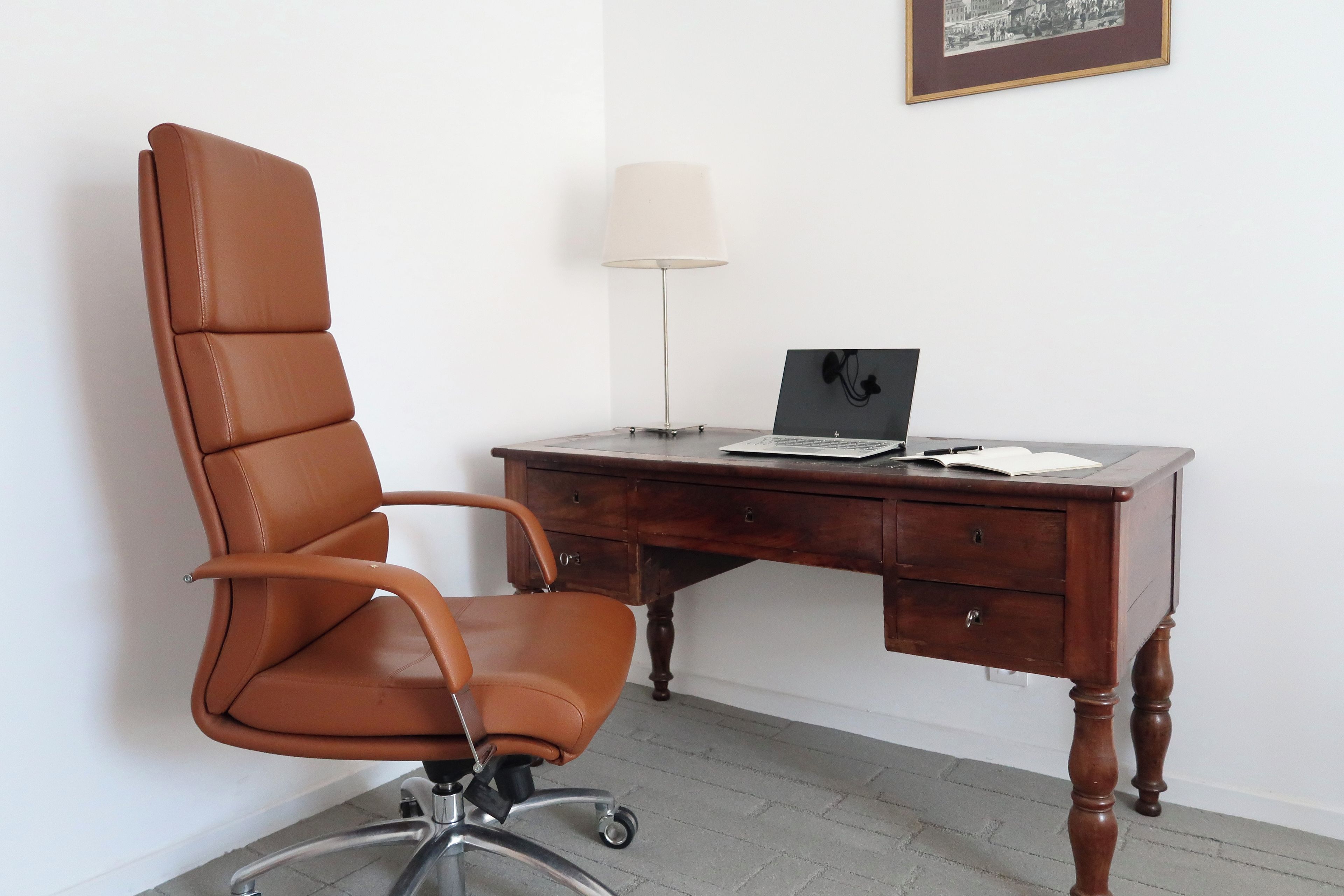 Una silla de oficina de cuero marrón con ruedas, un escritorio de madera con una computadora portátil, una lámpara y un cuadro en la pared.
