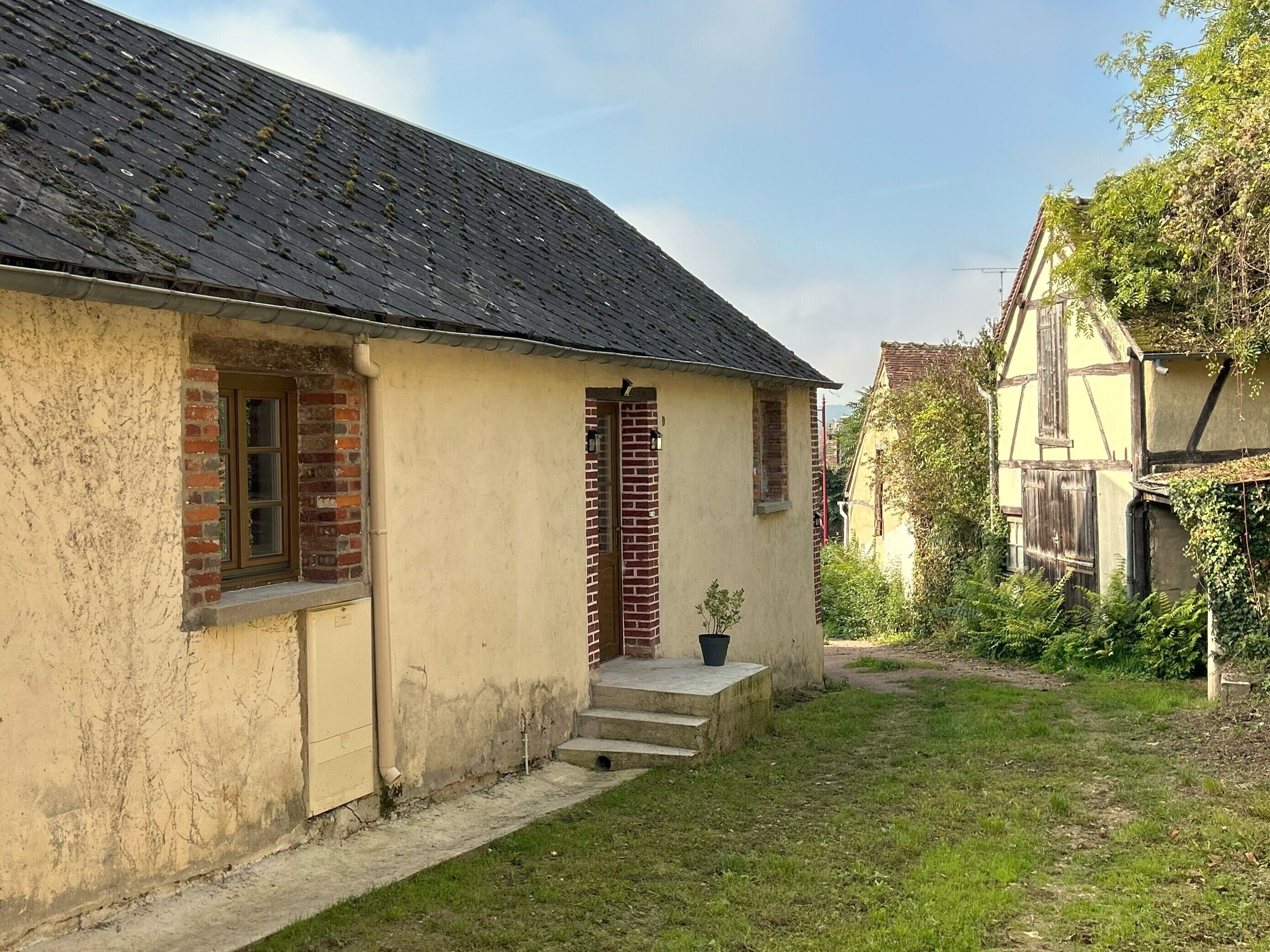 Een klein, laag huis met een schuin dak en een bakstenen schoorsteen. Er is een kleine veranda met treden die naar de deur leiden, en een bloempot op de treden.