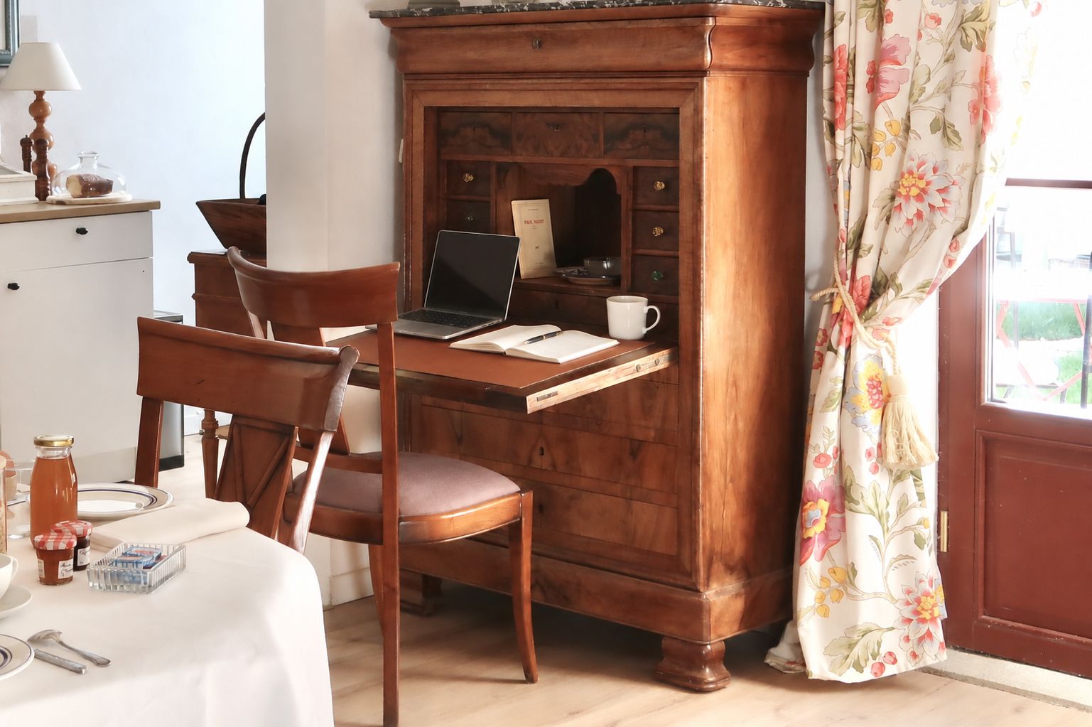 A wooden desk with a laptop, a chair, a small cabinet, and a window with floral curtains.