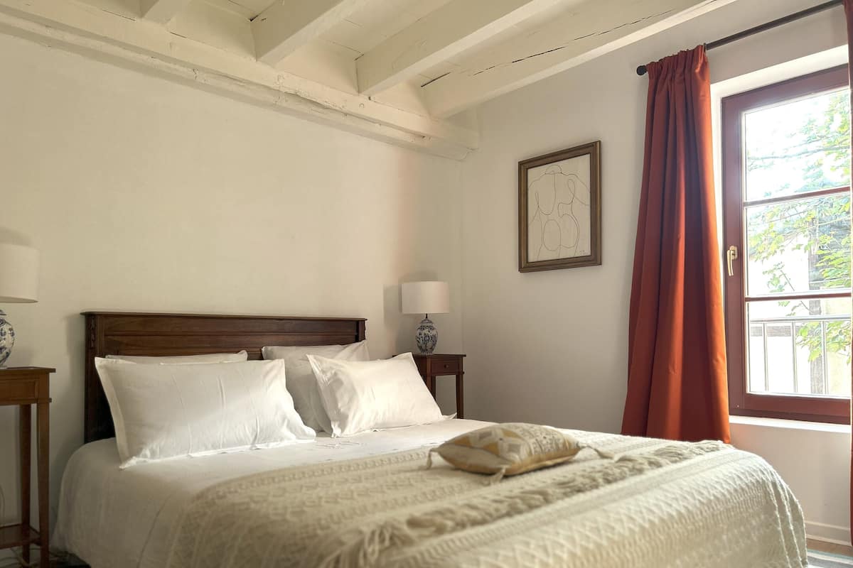 Een slaapkamer met een houten hoofdeinde, wit beddengoed, een nachtkastje met een lamp, een ingelijst schilderij aan de muur en een raam met gordijnen.
