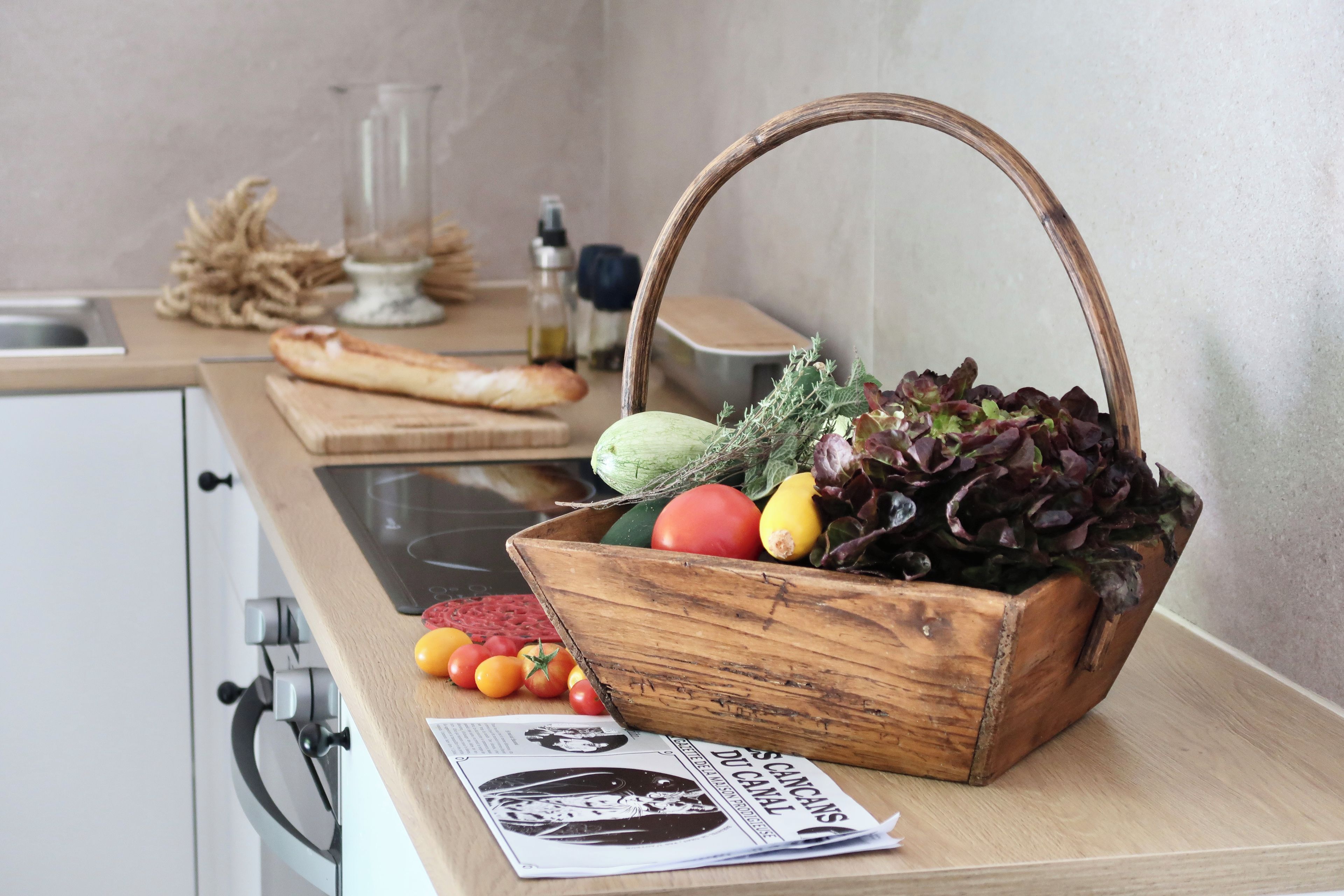 Una encimera de cocina con una cesta de madera llena de verduras, una tabla de cortar con pan y un periódico.