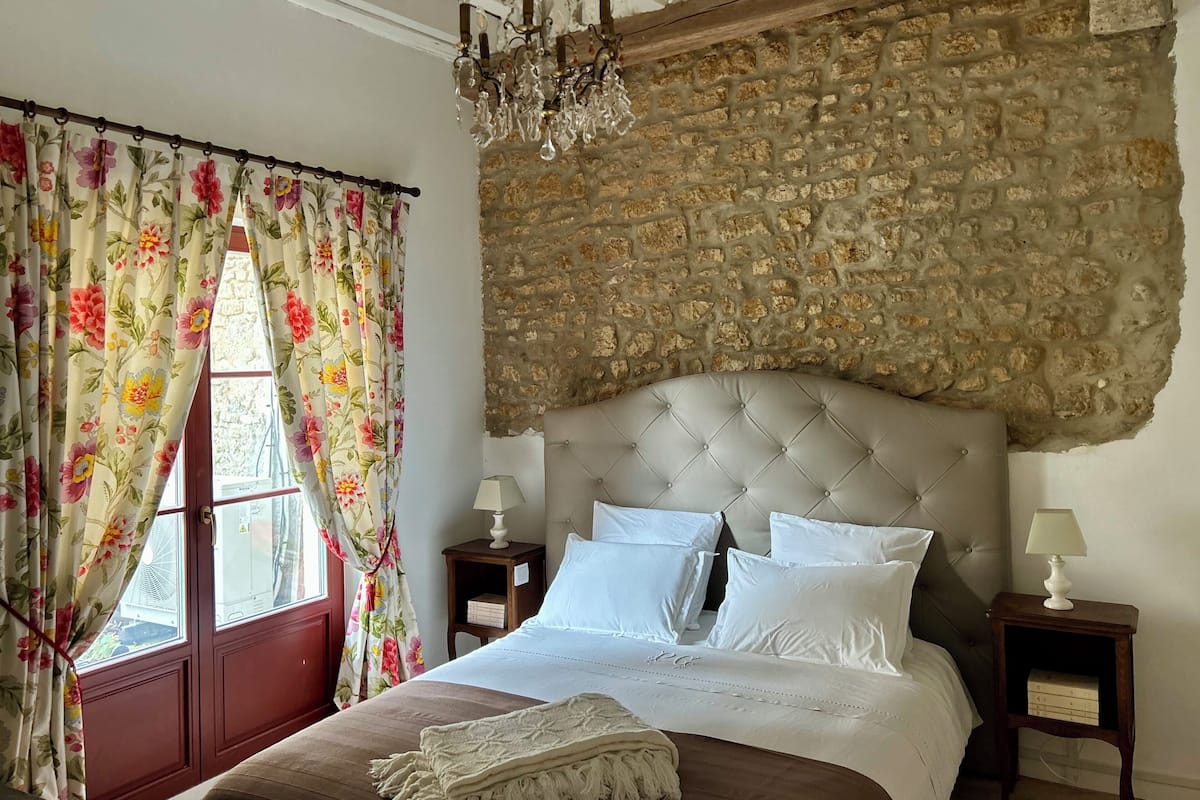 Een slaapkamer met een stenen muur, een gestoffeerd hoofdeinde, bloemen gordijnen en een kroonluchter.