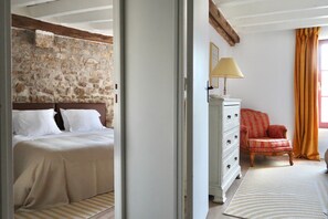 Une chambre avec un mur en pierre, un lit avec une literie blanche, une commode blanche avec des tiroirs, une chaise rouge et une fenêtre avec des rideaux.