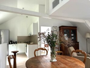A dining area with a wooden table, chairs, and a vase of dried flowers. A kitchen is visible in the background with a cabinet and a refrigerator.