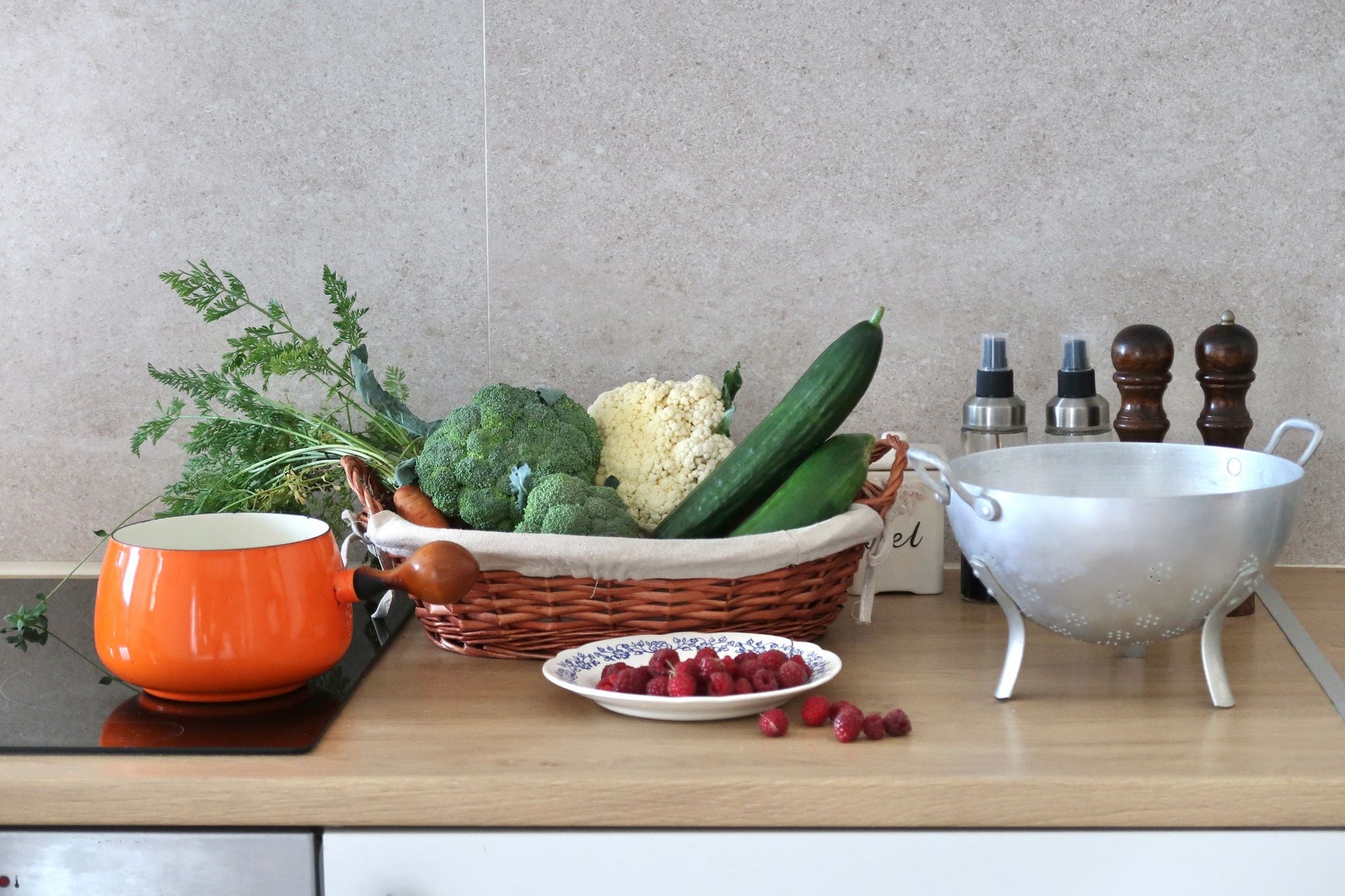 Una encimera de cocina con una cesta de verduras, un tazón de frambuesas y un colador.
