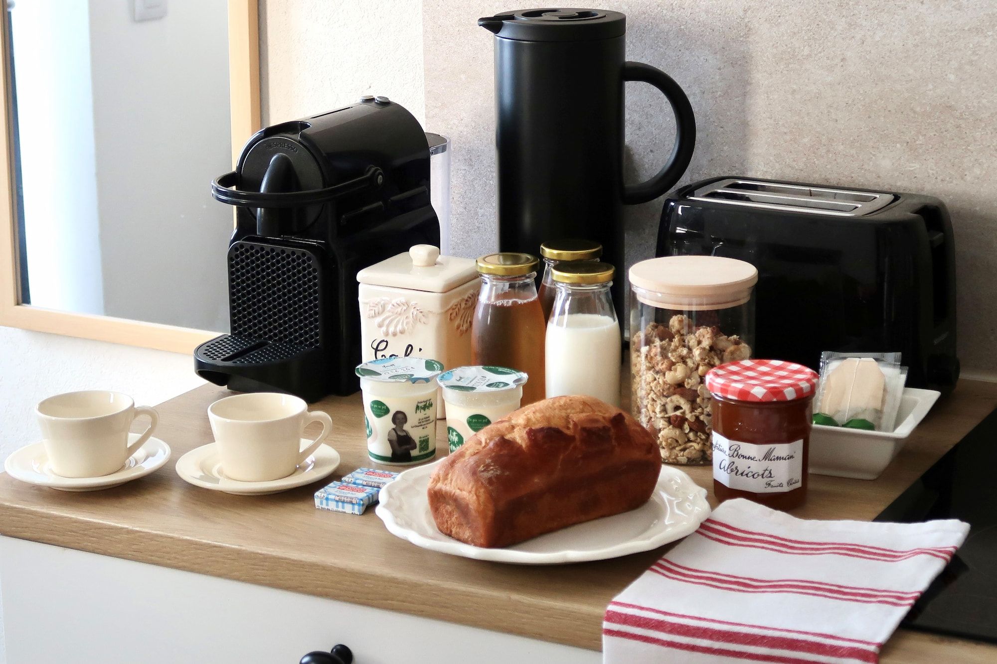 Preparación para el desayuno con cafetera, tostadora, leche, yogur, pan y mermelada.