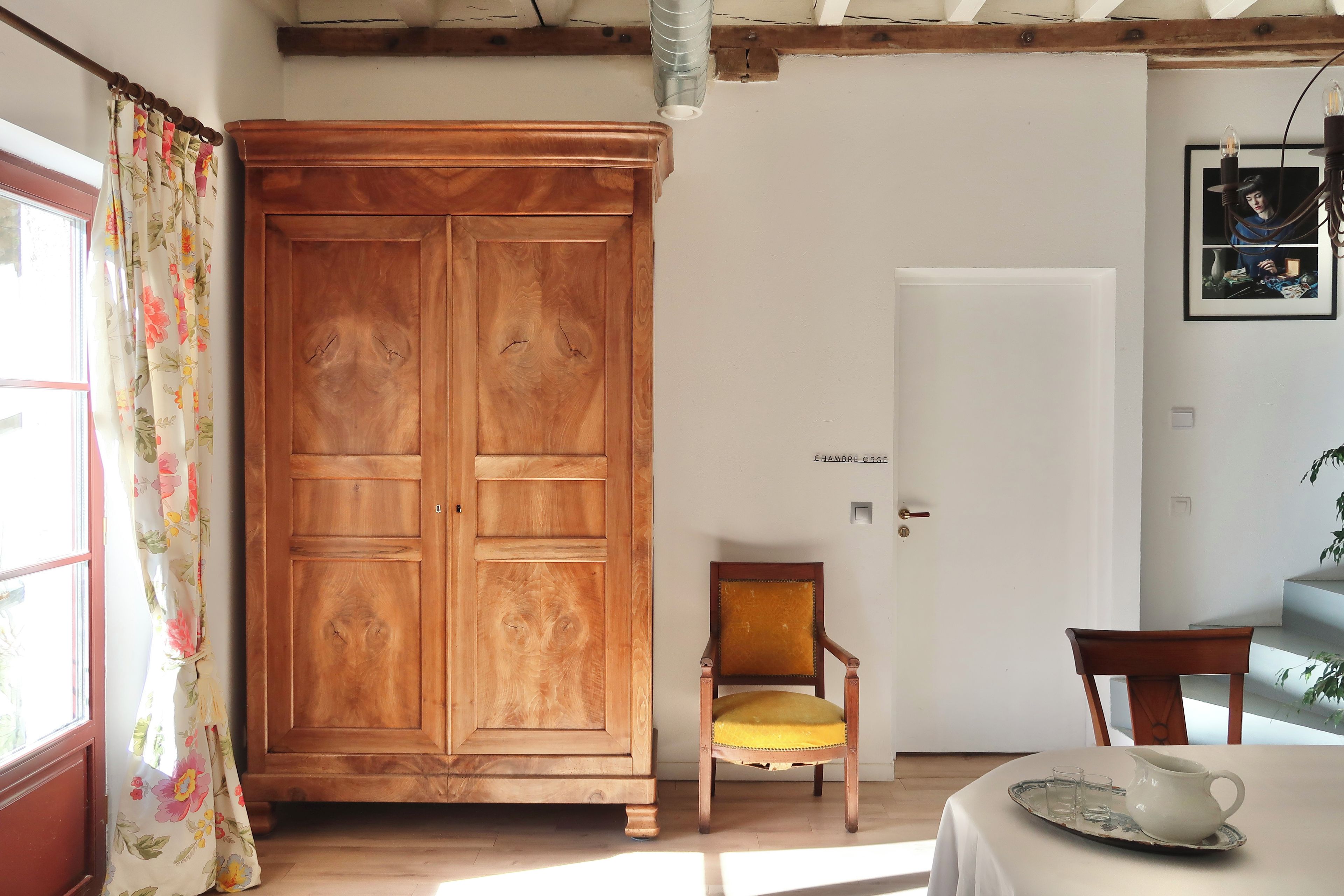 Una habitación con un armario de madera, una silla con cojín, una mesa con jarras y vasos, y una puerta con interruptor de luz.