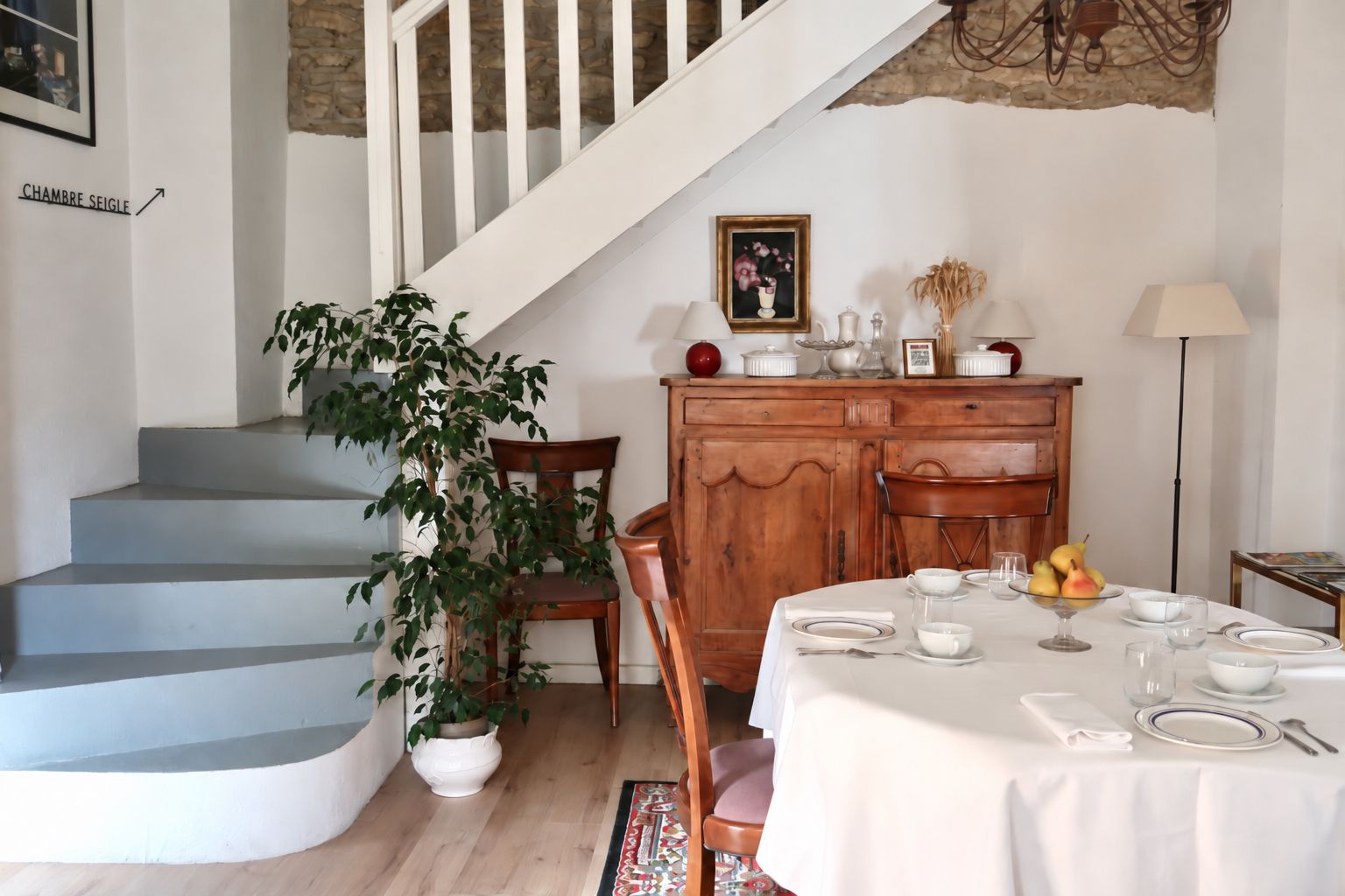 A dining area with a table set for four, a staircase, a wooden cabinet, and a potted plant.
