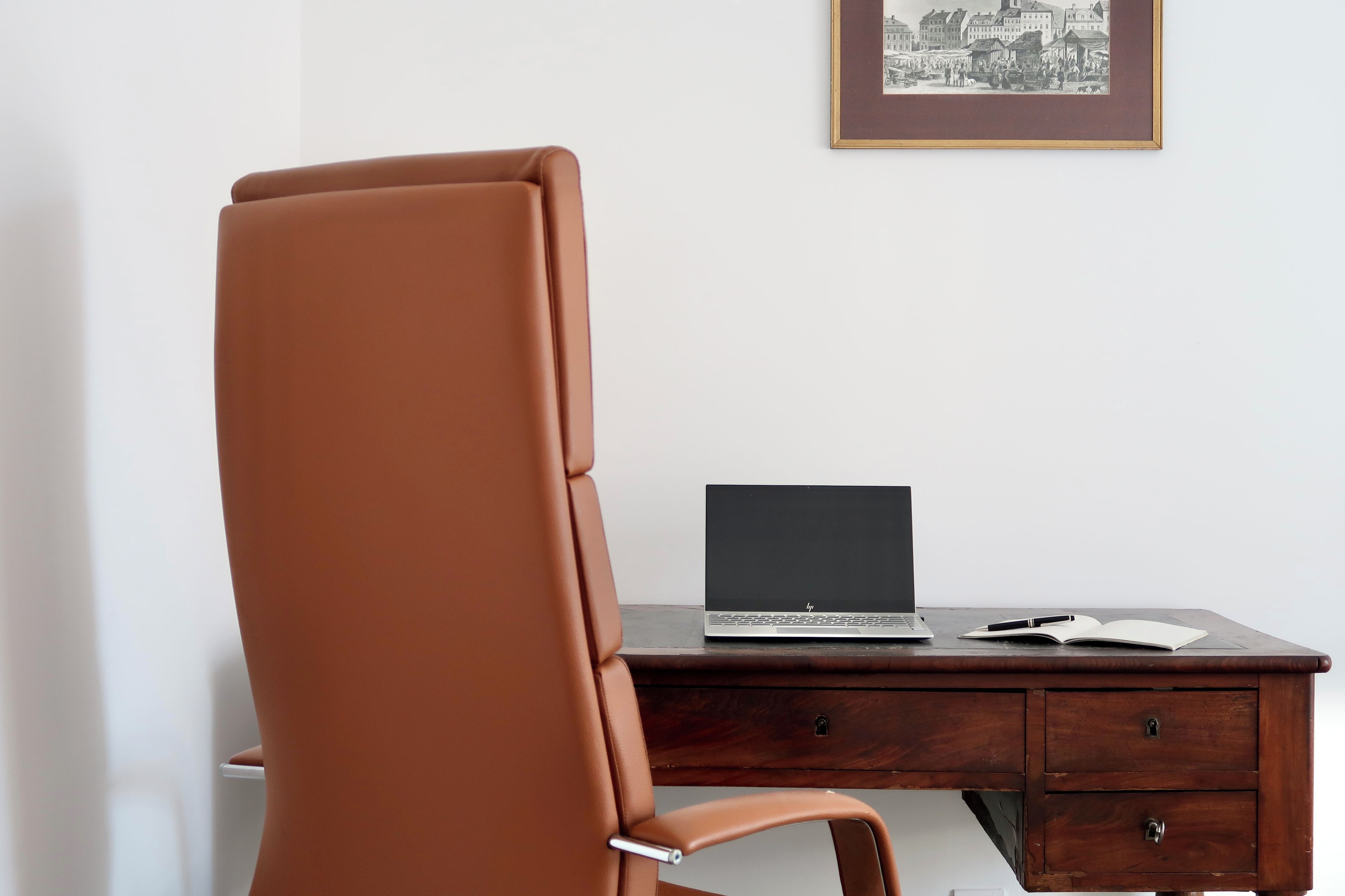 Una silla de cuero marrón, un escritorio de madera con una computadora portátil y un cuadro en la pared.