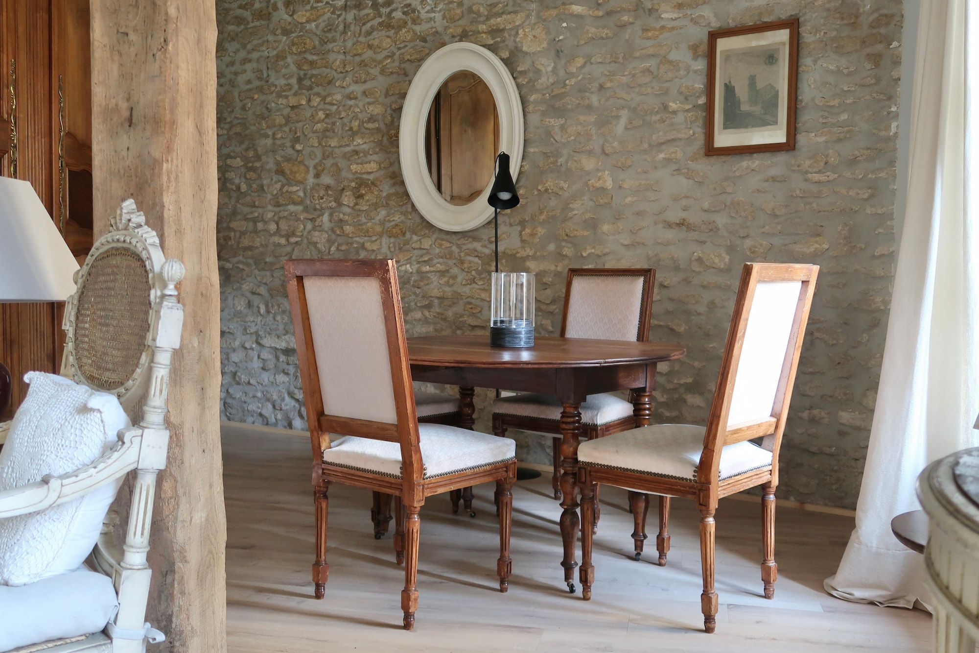 A dining area with a round wooden table, four chairs, a mirror, and a framed picture on a stone wall.